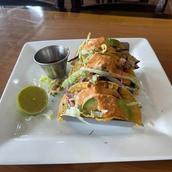 Three tacos with orange sauce, lettuce, and avocado, served with a lime wedge and dipping sauce.