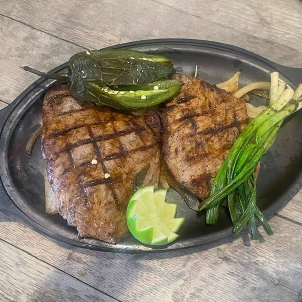 Two grilled steaks with charred peppers, green onions, and a lime wedge on a dark plate.