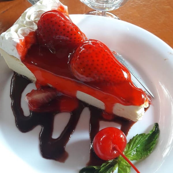 A slice of cheesecake topped with strawberries, strawberry sauce, whipped cream, chocolate drizzle, and a cherry.